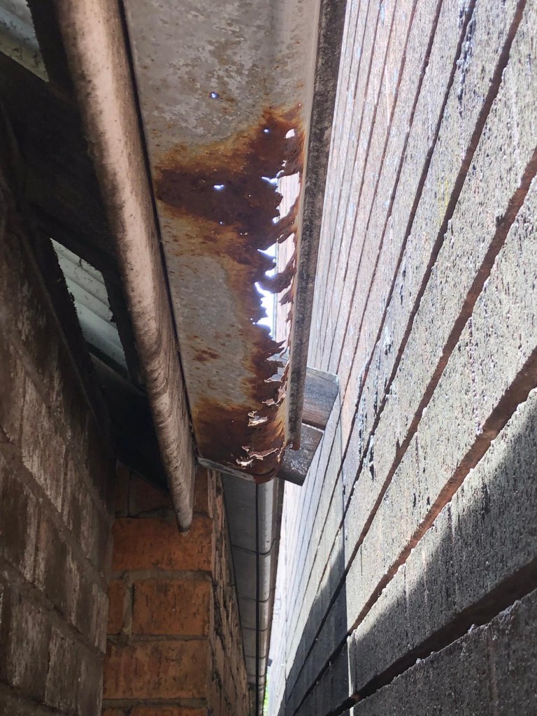 Rusted roof gutter with holes draining water into the narrow gap between adjacent brick buildings