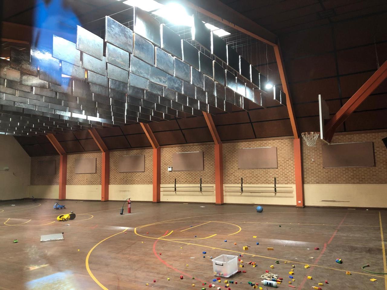 Empty gymnasium with a basketball hoop, scattered toys, and a plastic container on the floor; sunlight streams in from windows near the ceiling.